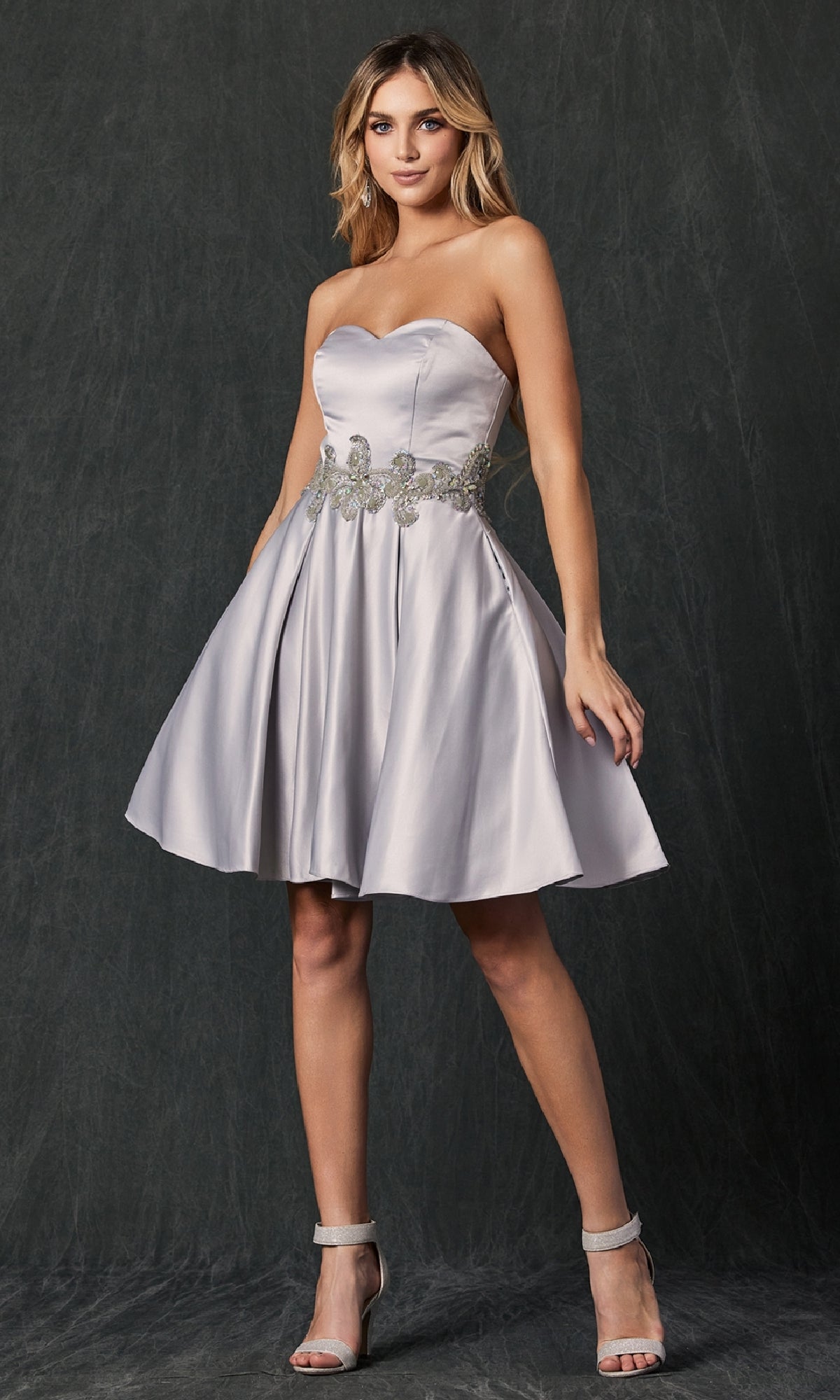 A woman poses before a dark backdrop in the Juliet 769 Short Hoco Dress, a strapless silver knee-length design featuring subtle beading, a decorative belt, and silver high-heeled sandals for extra shimmer.